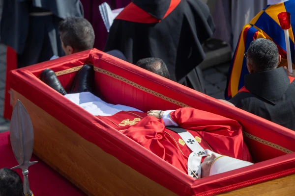 The body of Pope Francis, vested in traditional red papal funeral vestments, lies in state in a simple wooden coffin lined with red silk on April 23, 2025. The late pontiff's hands are folded in prayer, holding a rosary. Credit: Daniel Ibáñez/CNA
